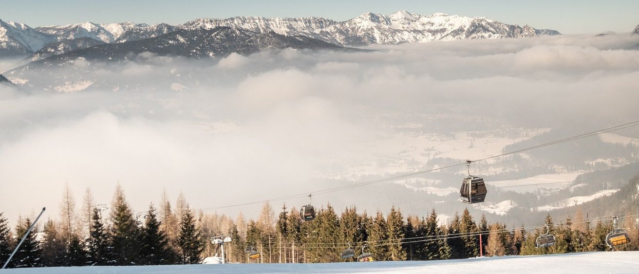 Eine verschneite Berglandschaft mit einem Seilbahnsystem im Vordergrund, verschneiten Bäumen und mit Nebel bedeckten Bergen.