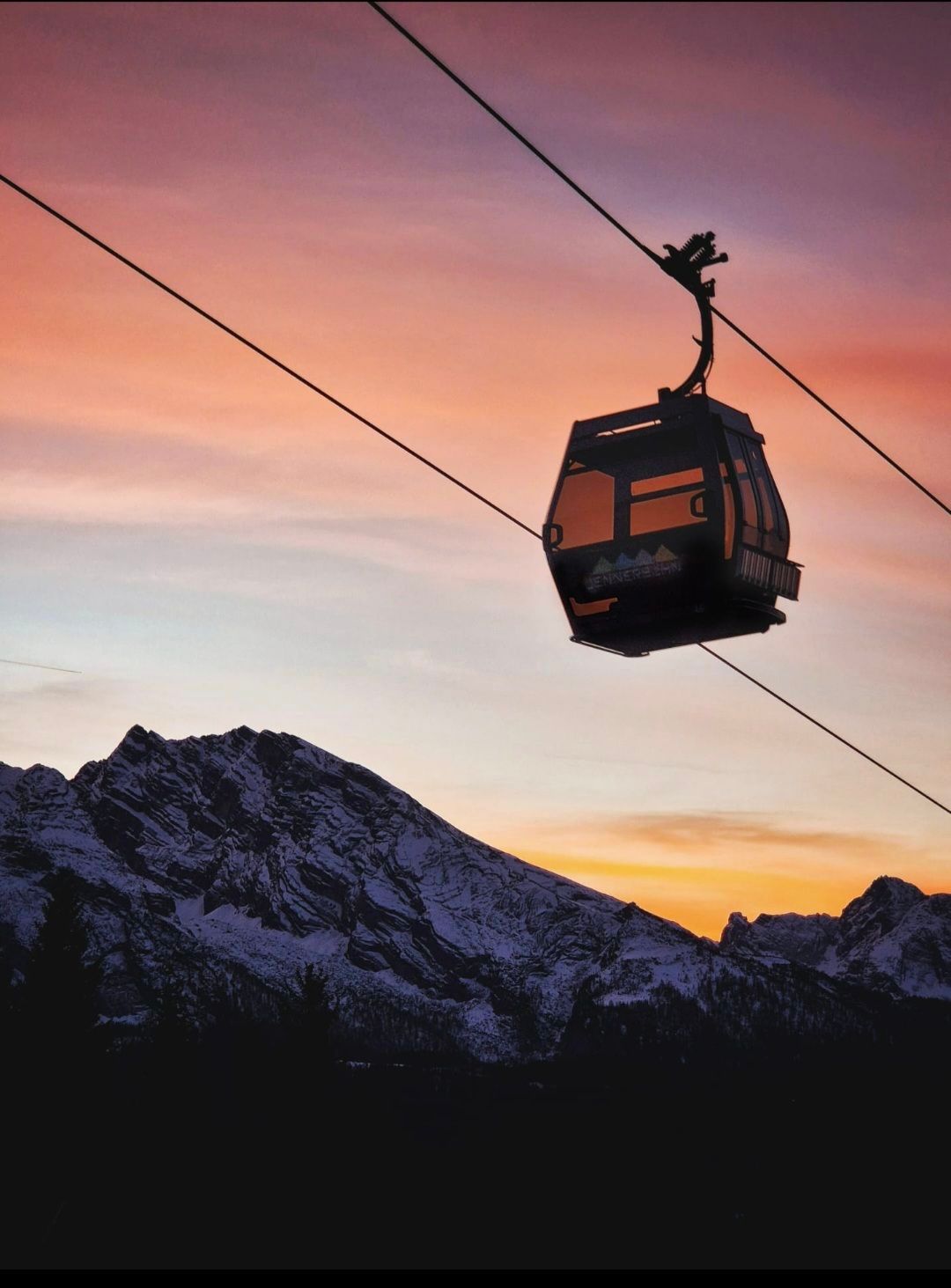 Eine Seilbahn, die von Drähten gehalten wird, vor dem Hintergrund eines verschneiten Gebirgszugs bei Sonnenuntergang, mit einem lebhaften orangefarbenen und rosafarbenen Himmel.