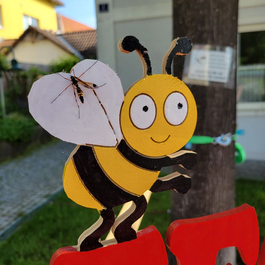 Eine hölzerne Bienenfigur auf einem roten Schild, mit einem Baum und Gebäuden im Hintergrund.