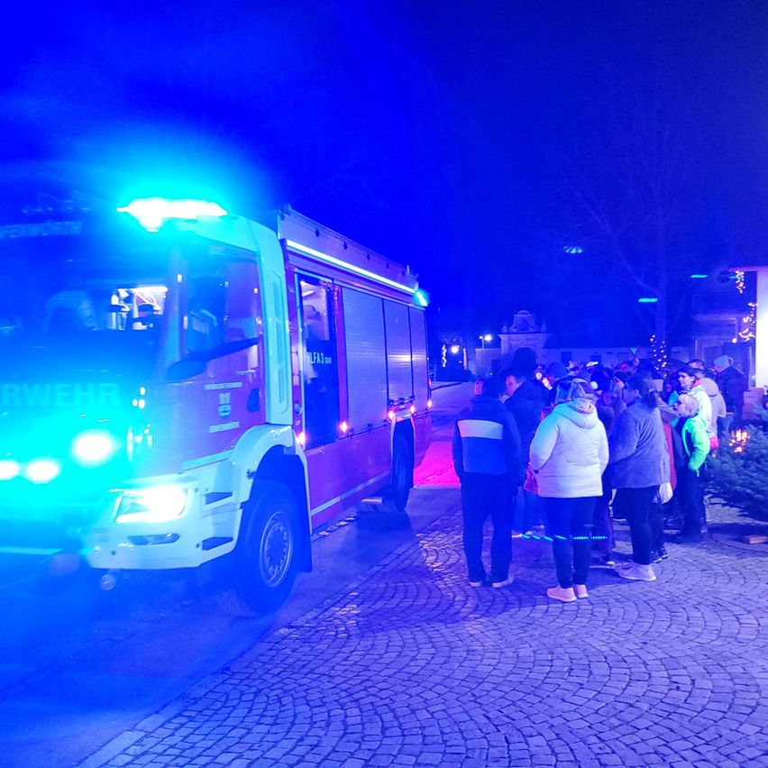 Ein Feuerwehrauto mit blauen Lichtern steht nachts auf einer Kopfsteinpflasterstraße. Mehrere Menschen stehen vor dem Auto.