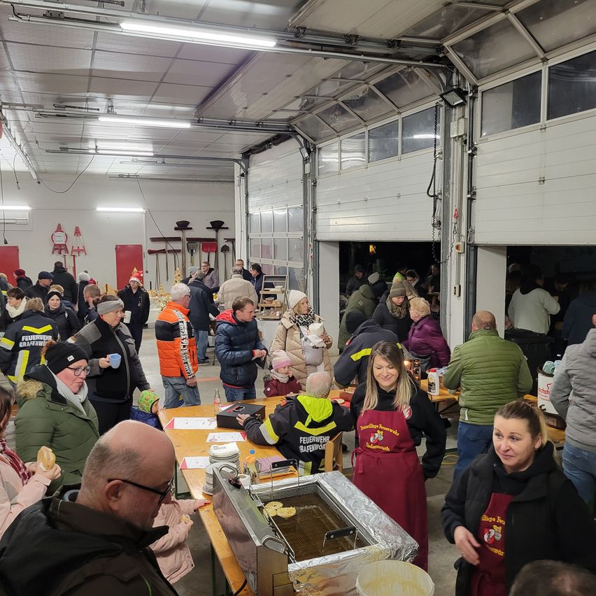 Eine große Menschenmenge in Winterkleidung steht in einem Raum mit mehreren Tischen, einige essen. Der Raum hat eine hohe Decke mit Lichtern und zwei offene Garagentore.
