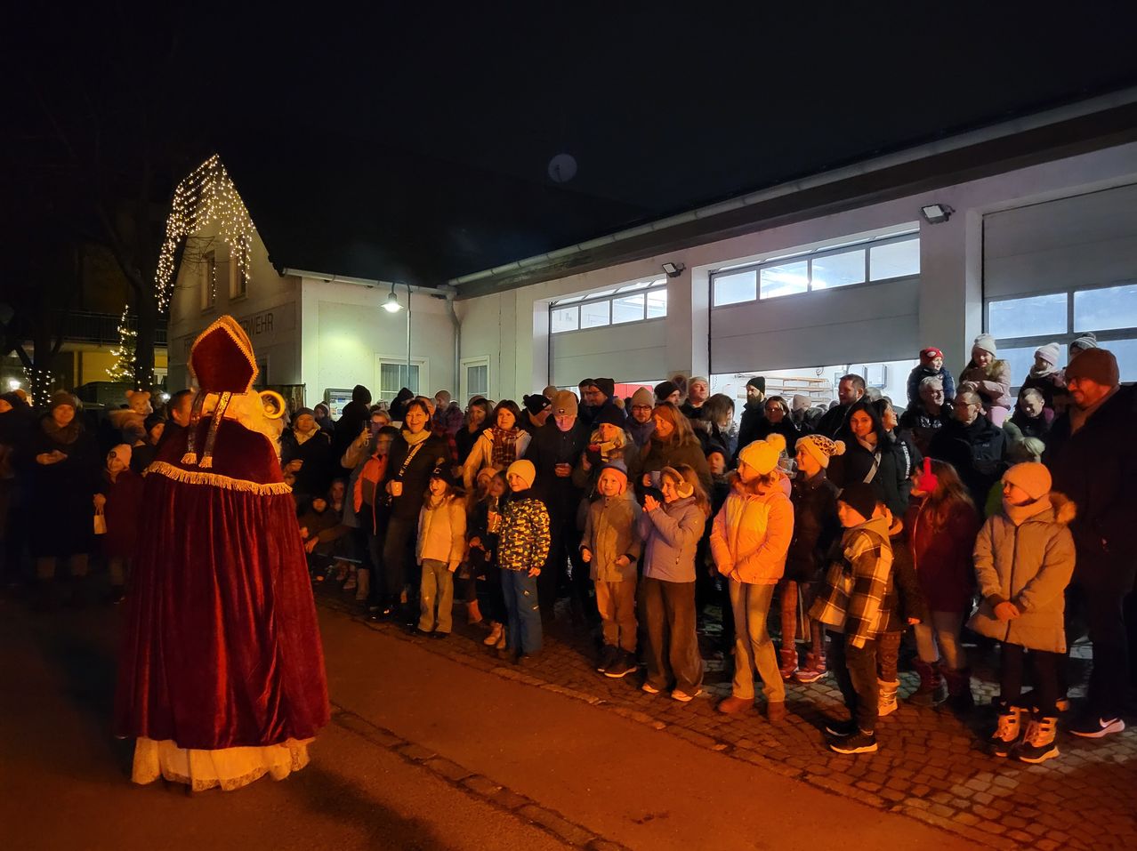 In der Nacht versammelt sich eine Menschenmenge auf einer Kopfsteinpflasterstraße und beobachtet einen Mann in einem roten Gewand und Hut. Sie stehen vor einem Gebäude mit Lichtern und Glasfenstern.
