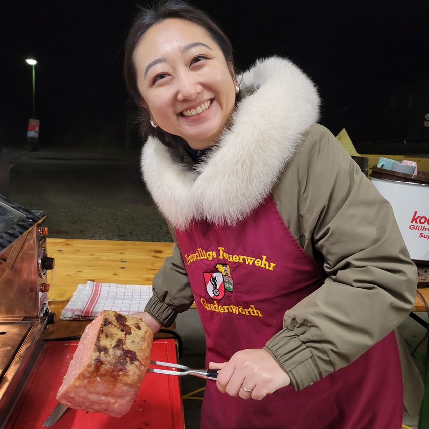 Eine Frau in einem mit Fell gefütterten Mantel und einer Schürze steht im Freien, lächelt und hält eine Gabel mit einem großen Stück Fleisch. Sie steht vor einem Grill und einem Holztisch. Der Hintergrund ist dunkel, mit einer Straßenlaterne und Lagerboxen.