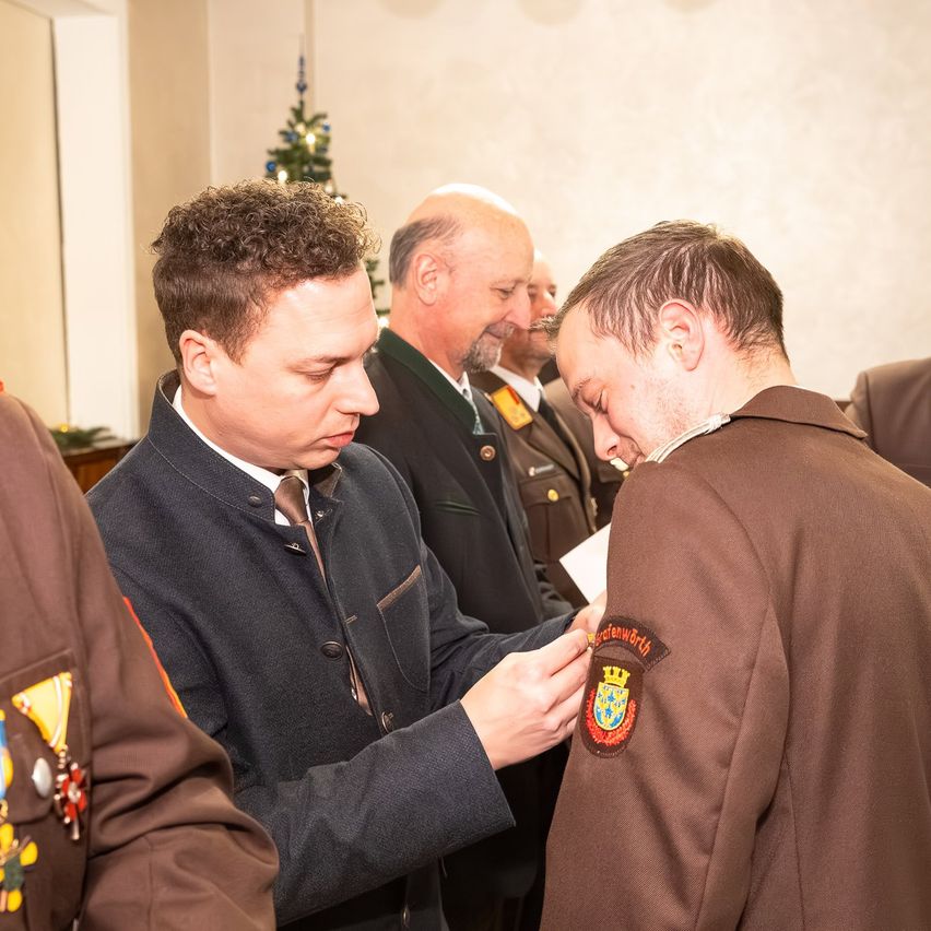 Ein Mann in einem Anzug befestigt eine Abzeichen an der Uniform eines anderen Mannes bei einer formellen Veranstaltung, mit einem Weihnachtsbaum im Hintergrund.
