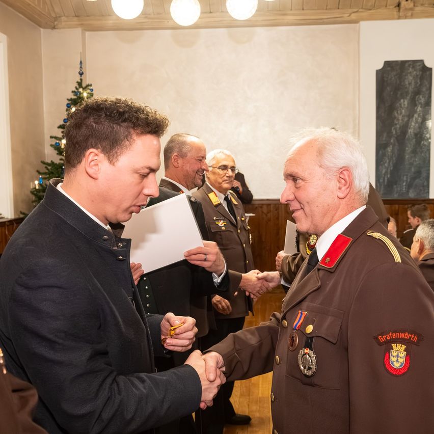 Ein älterer Mann in Uniform schüttelt einem jüngeren Mann die Hand in einem Saal. Dahinter hält ein Mann in Anzug ein Papier. Eine Weihnachtsbaumecke steht in der Ecke.