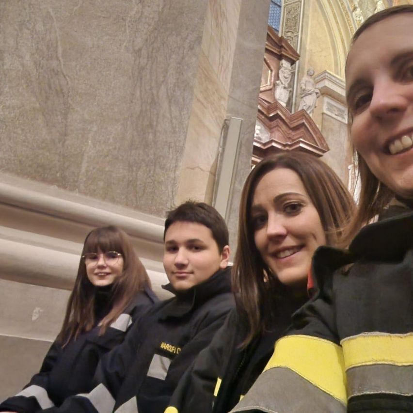 Vier Menschen lächeln in einer Kathedrale. Zwei tragen schwarze und gelbe Uniformen, und zwei tragen schwarze Jacken. Sie stehen vor einer Steinmauer.
