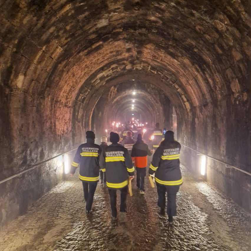 Feuerwehrleute gehen durch einen schwach beleuchteten Tunnel mit gewölbten Decken, tragen Uniformen und Ausrüstung. Der Tunnel hat Steinwände und Kopfsteinpflaster.