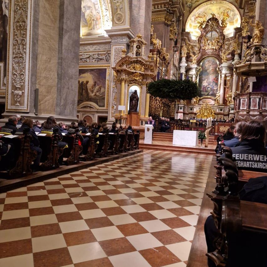 Eine Gruppe von Feuerwehrleuten sitzt in einer Kirche und nimmt an einer Zeremonie teil. Die Kirche ist mit aufwendigen Verzierungen geschmückt, und die Kirchenbänke sind kariert. Im Vordergrund steht ein Podest.