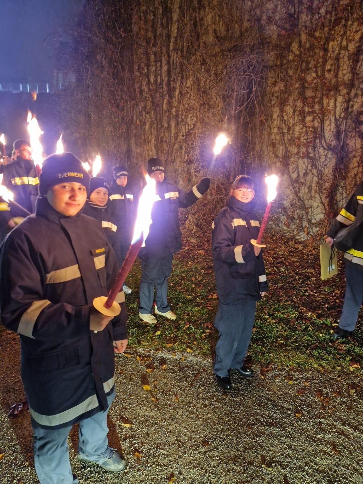 Eine Gruppe von Feuerwehrleuten mit brennenden Fackeln steht bei Nacht draußen, mit einer von Efeu bedeckten Wand im Hintergrund.