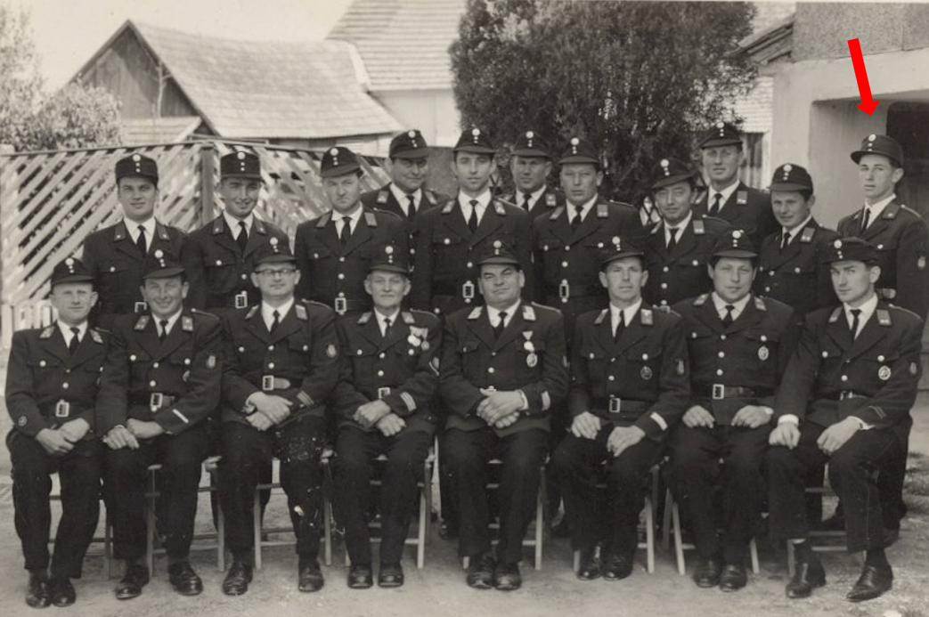 Ein altes Schwarz-Weiß-Foto zeigt eine Gruppe uniformierter Männer, möglicherweise Polizisten, die auf Stühlen sitzen. Sie tragen Mützen und Krawatten. Der Hintergrund zeigt ein Gebäude mit Dach und einem Baum.