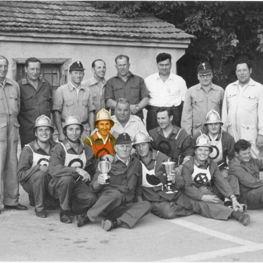 Eine Gruppenaufnahme von Männern in Uniformen und Helmen, die Trophäen halten, stehend und sitzend vor einem Gebäude.