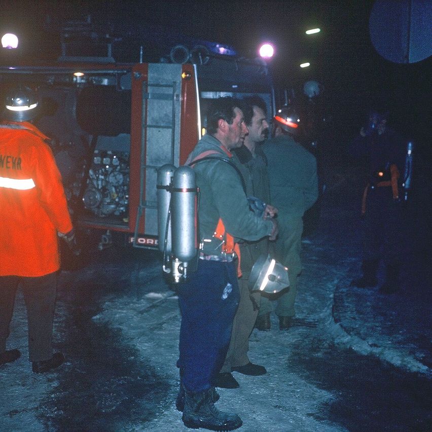 Eine Gruppe von Feuerwehrleuten in Schutzausrüstung steht nachts bei einem Feuerwehrwagen. Sie scheinen sich auf eine Mission vorzubereiten.