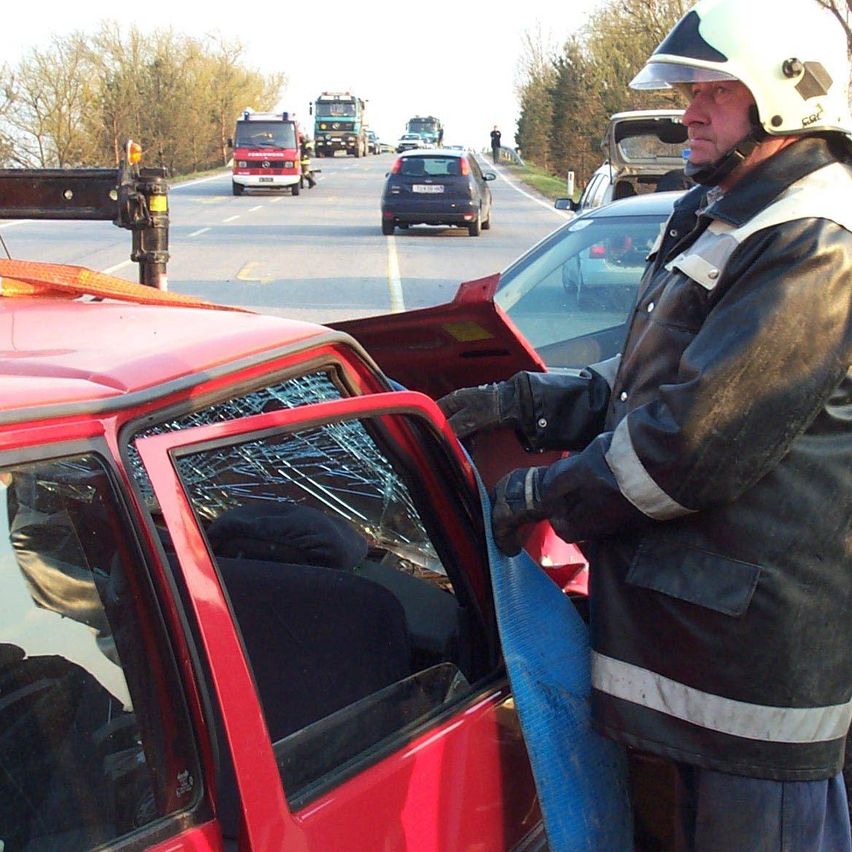 Ein Feuerwehrmann in einem Helm steht neben einem demolierten roten Auto am Straßenrand. Eine Person sitzt im Auto. Andere Fahrzeuge bewegen sich auf der Straße.