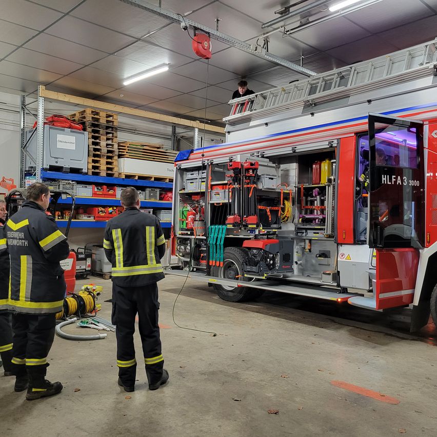 Zwei Feuerwehrleute stehen in einer Garage mit einem Feuerwehrauto. Die Türen des Fahrzeugs sind geöffnet, und eine Person steht auf der Leiter. Lagerboxen stehen auf den Regalen, und ein Feuerlöscher ist auf dem Boden abgestellt.