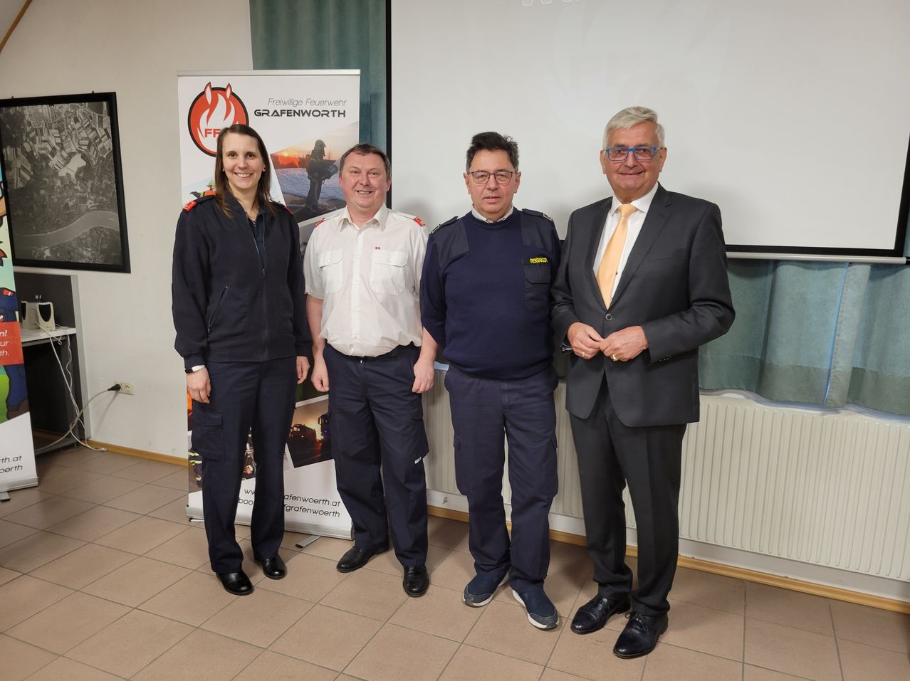 Vier Personen in Uniformen stehen lächelnd vor einer weißen Wand für ein Foto. Sie befinden sich in einem Raum mit gefliesten Boden.