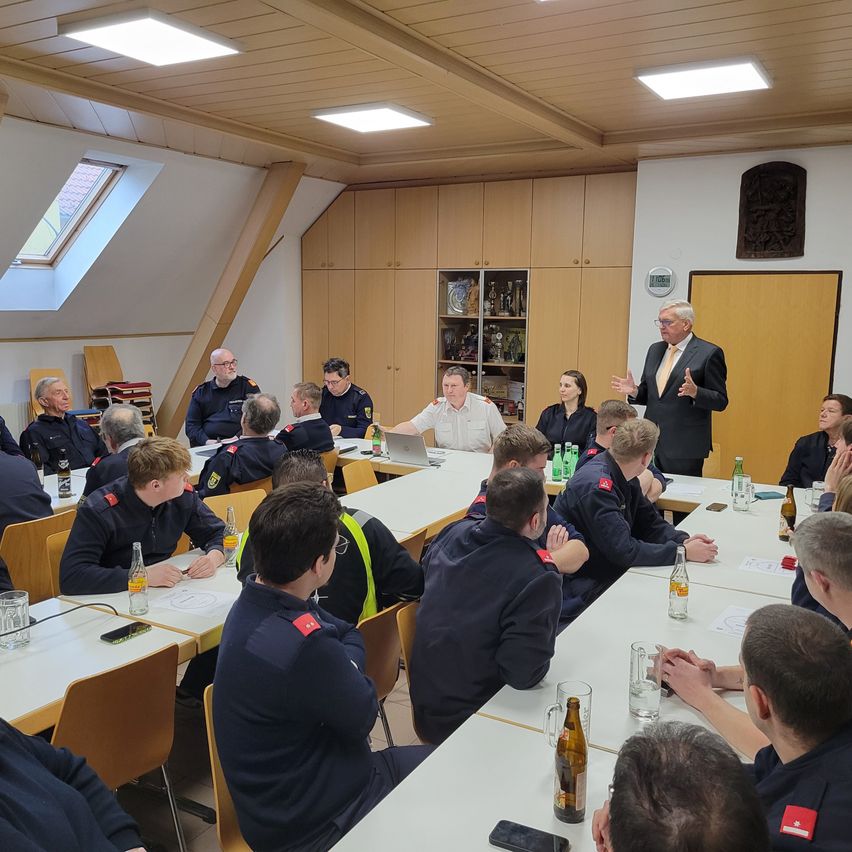 Eine Gruppe von Menschen in Uniformen sitzt an Tischen in einem Raum, vor ihnen steht ein Mann. Auf den Tischen stehen Flaschen, Tassen und Papiere. Holzschränke und Regale stehen an der Wand. An der Wand sind eine Uhr und ein Bilderrahmen angebracht.