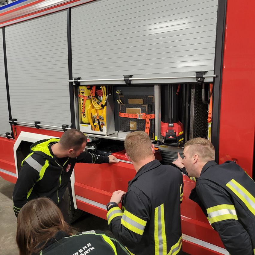 Four firefighters examine equipment in a fire truck. A man points to the open compartment. Another points to the equipment inside.