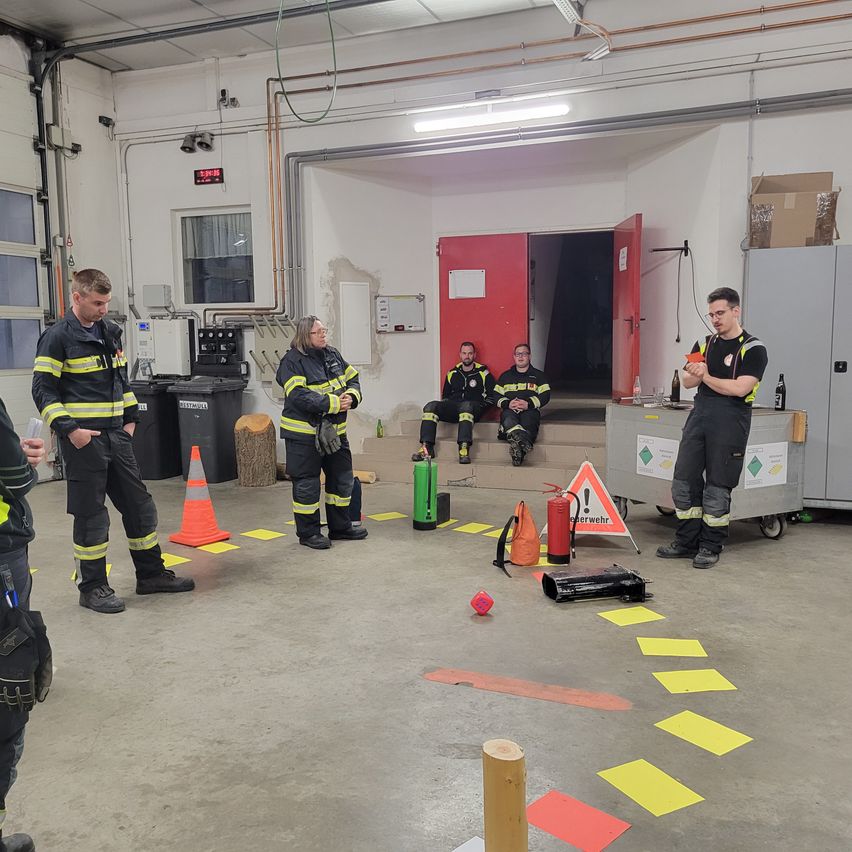 Firefighters in a training session, standing and sitting around an open red door, with safety cones, a fire extinguisher, and a yellow floor layout.