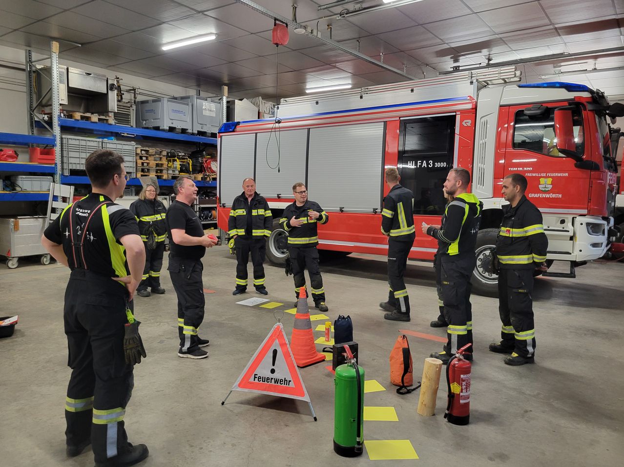 A group of firefighters stand around a firetruck inside a garage. They are wearing uniforms and gloves. They are discussing something. There is a fire extinguisher, a caution triangle, and a cone on the floor.