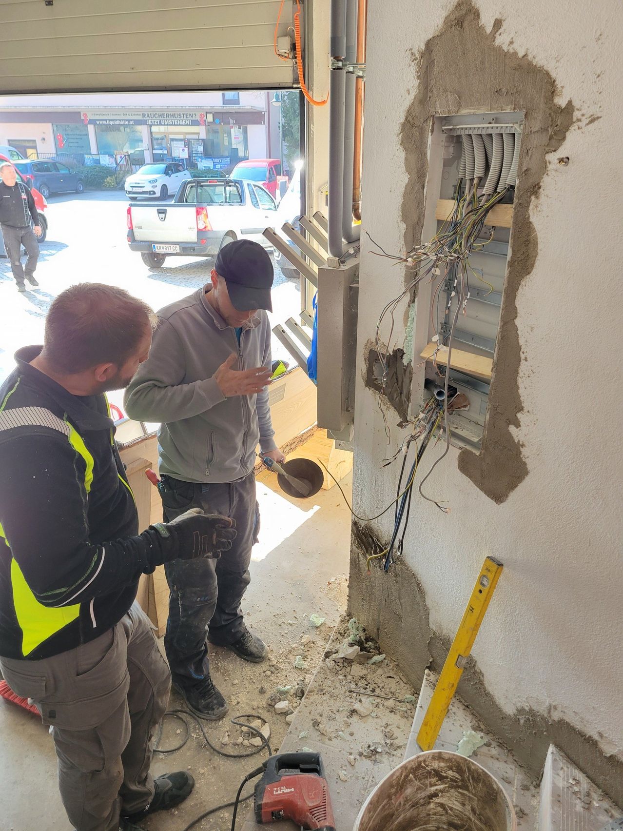 Two men are working on the wall with wires in a building. One man is holding a tool. Another man is looking at the wall.