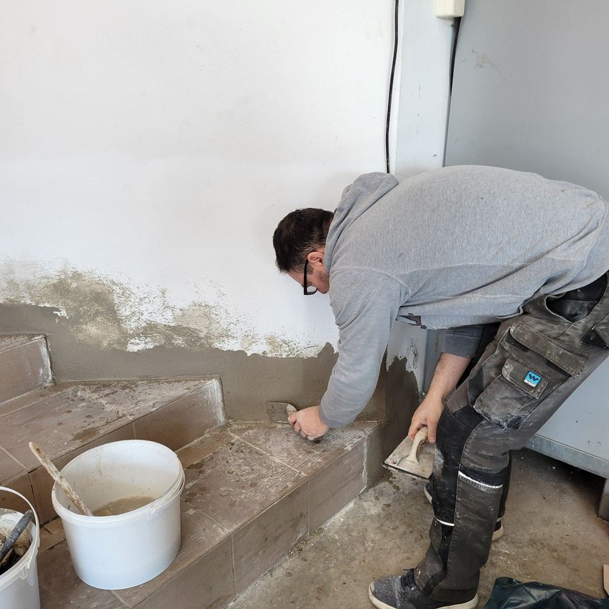 A man in a gray hoodie and black pants is applying cement to a step. A white bucket with a wooden stick is on the step.