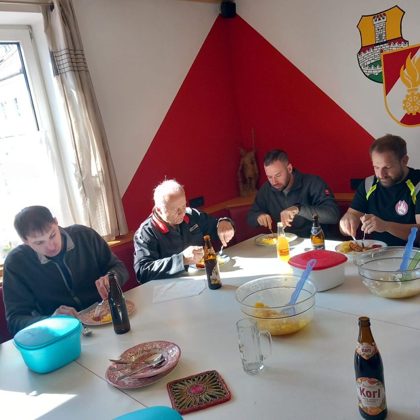 Five men sit at a table, enjoying a meal. A window with curtains is behind them, and a wall with a logo is visible. Various bottles, plates, and bowls are on the table.