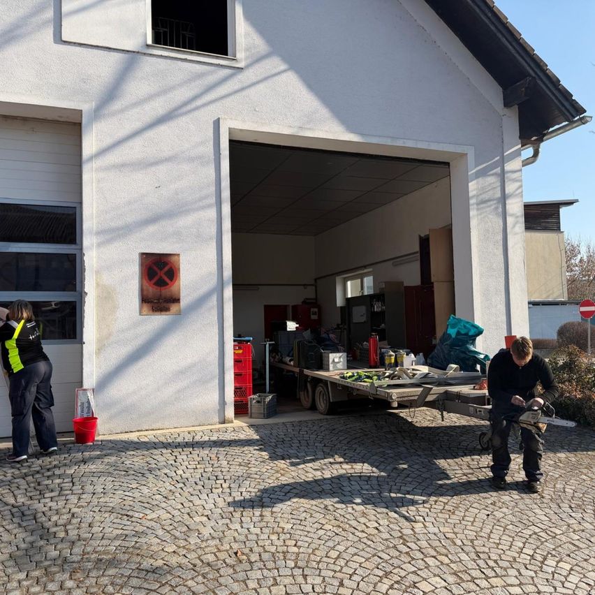 Two people work outside a building with a sign. One holds a red bucket and the other has a chainsaw.