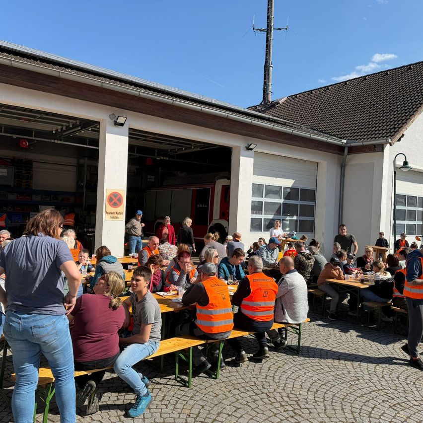 A group of people are sitting at tables outside a building. They are wearing orange vests. The building has garage doors. There is a sign on the wall with a red cross.