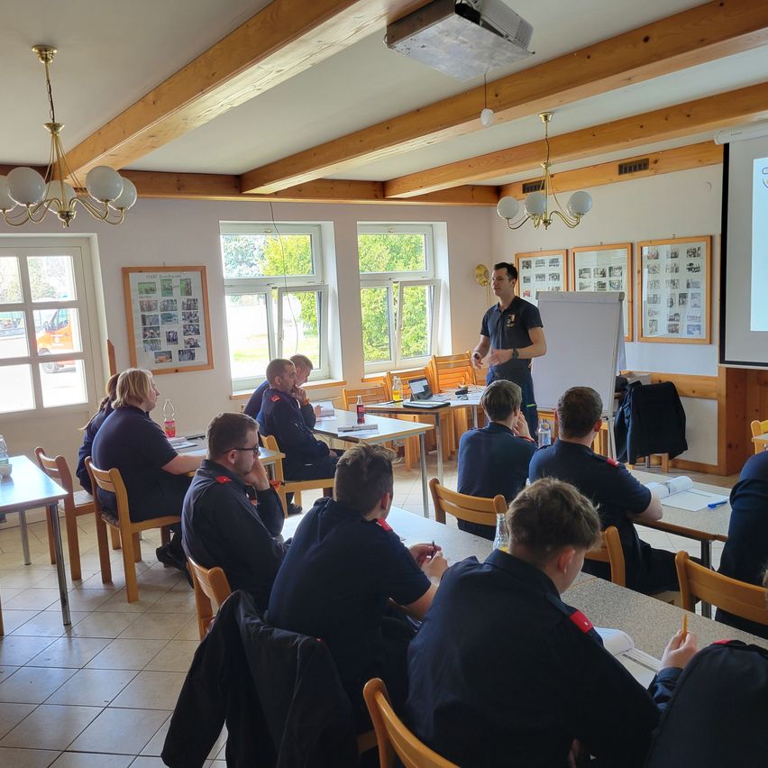 A classroom with wooden beams and chandeliers has a group of students listening to a teacher. The room has desks, chairs, and a whiteboard. Some students are looking at papers, while others are looking at the teacher. There are also windows and picture frames on the wall.