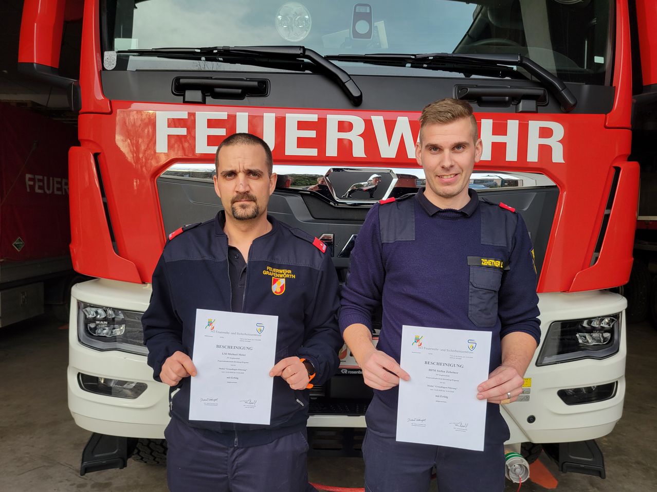 Two men stand in front of a red fire truck, holding certificates. Both wear uniforms, one with a logo on the left chest and the other with a logo on the right. They are smiling.