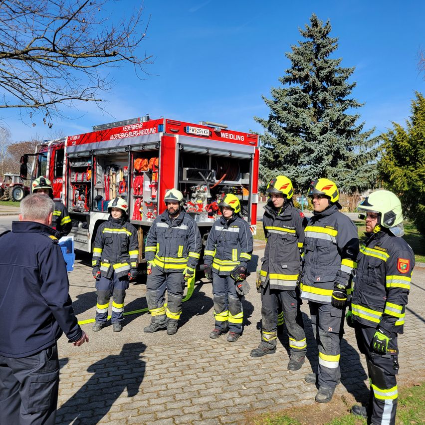 A group of firefighters in full gear stand in front of a red fire truck with an open compartment on a sunny day.