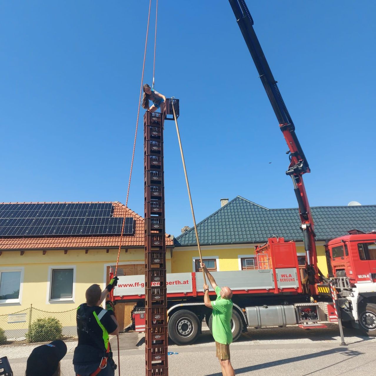 Eine Frau wird von einem Kran mit zwei Männern, die Seile halten, angehoben. Der Kran ist an einem Lkw befestigt, auf dem 'WLA' steht. Das Gebäude dahinter hat Solarmodule auf dem Dach.