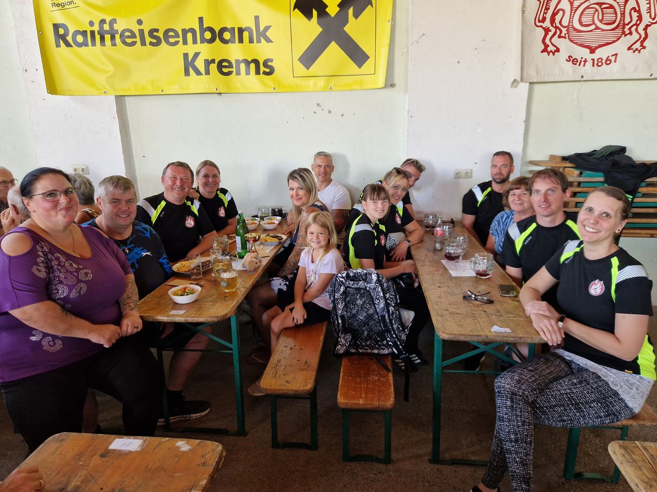 A group of people sitting at a long table in a restaurant, some wearing black and yellow vests. A girl sits on a bench in front of a backpack. A banner with the text 'Eisenbank Krems' hangs above.