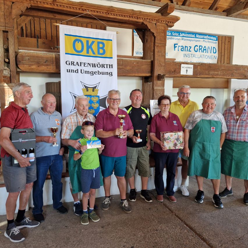A group of elderly people and a young boy stand together in front of a banner, holding trophies and gifts. They are all smiling and appear to be celebrating an achievement.