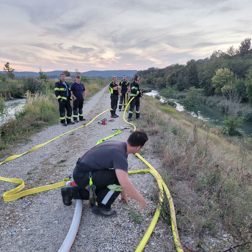 Firefighters are preparing hoses near a river, with one man kneeling and others standing by.