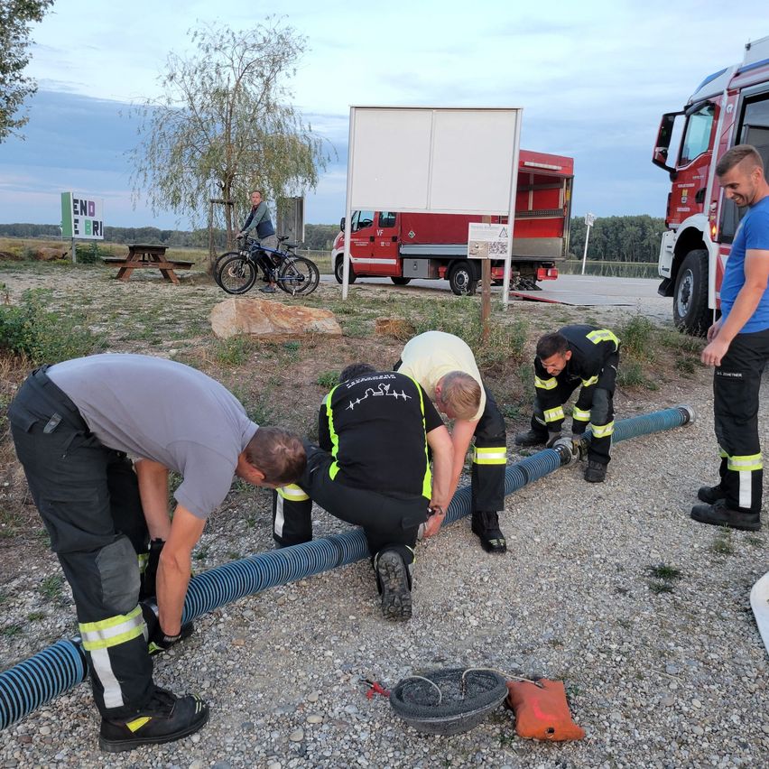 A group of firefighters are working on a long blue hose on a gravel surface. A fire truck is parked nearby, and a man is riding a bicycle.