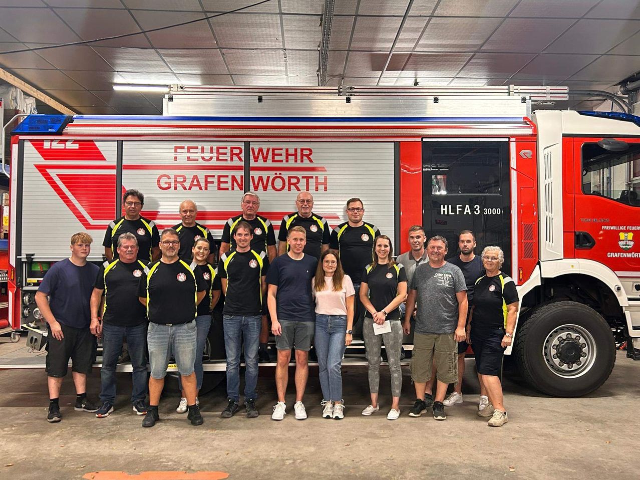 A group of people, including men and women, stand in front of a fire truck with the words 'Feuer Wehr Grafen Worth' on it. They are all smiling and appear to be posing for a photo.