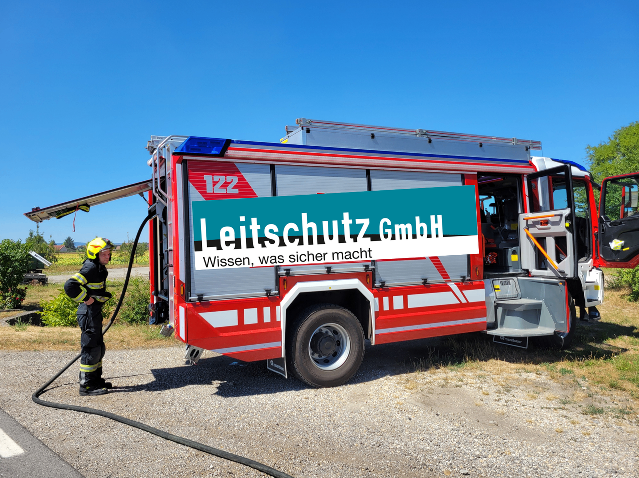A firefighter stands next to a red fire truck with a sign that reads 'Leitschutz GmbH' and 'Wissen, was sicher macht'. The truck is parked on gravel.