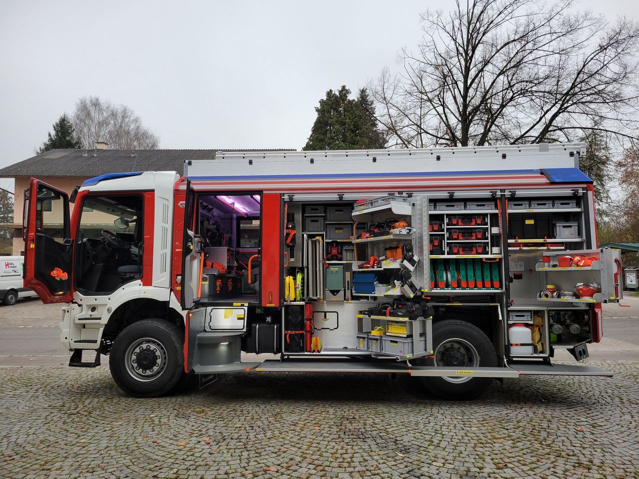Ein rot-weißer Feuerwehrwagen mit offenen Türen und Fächern voller Werkzeuge und Ausrüstung, auf Kopfsteinpflaster geparkt, mit Bäumen im Hintergrund.