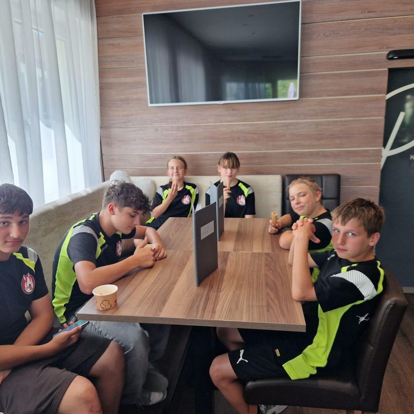 Sieben Jungen in Sporttrikots sitzen um einen Tisch und essen Eis und reden. Sie sind in einem Raum mit einer Holzwand und einem Fernseher.