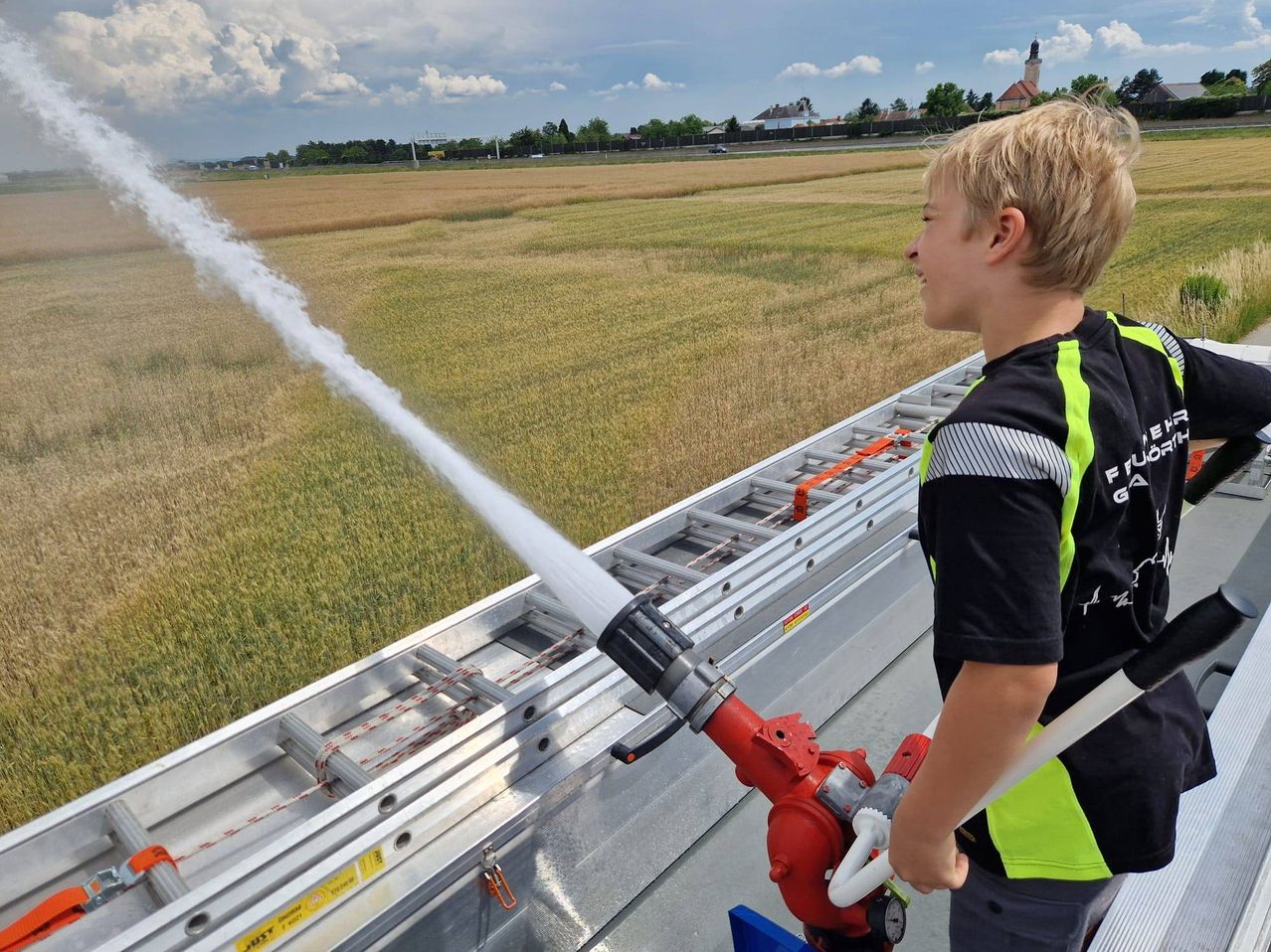 Ein junger Junge in einer reflektierenden Weste hält einen Feuerwehrschlauch und sprüht Wasser in die Luft auf einem großen Feld.