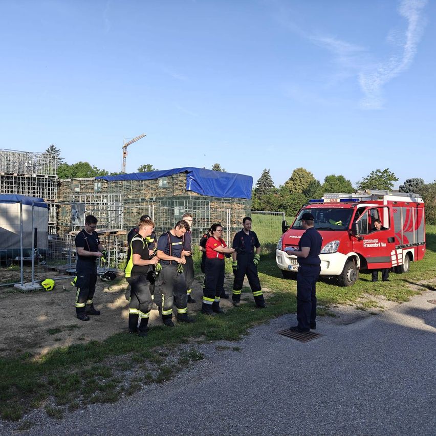 Eine Gruppe von Feuerwehrleuten steht bei einem roten Feuerwehrauto in einer Übung. Sie sind mit Sicherheitsausrüstung ausgestattet und besprechen etwas.