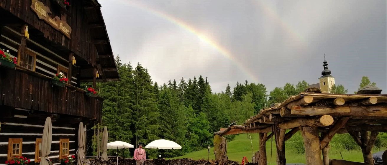 Ein Regenbogen erstreckt sich über einem Himmel über einem rustikalen Essbereich im Freien. Eine Person in einem rosa Shirt steht in der Nähe von Schirmen und Holztischen. Im Hintergrund sind Bäume und ein Kirchturm sichtbar.