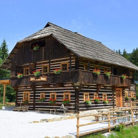 Ein traditionelles Holzhaus mit Strohdach. Rote Blumen in Töpfen schmücken die Fenster und Balkone. Ein Holzzaun umgibt das Haus.