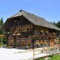 Ein traditionelles Holzhaus mit Strohdach. Rote Blumen in Töpfen schmücken die Fenster und Balkone. Ein Holzzaun umgibt das Haus.