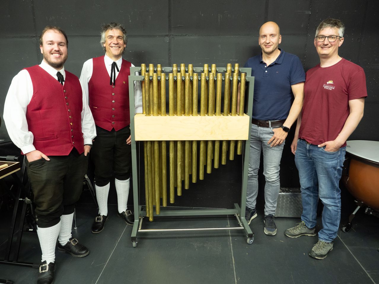 Vier Männer stehen neben einem metallischen Instrument, möglicherweise einer Glocke, auf einem Ständer. Sie alle lächeln.
