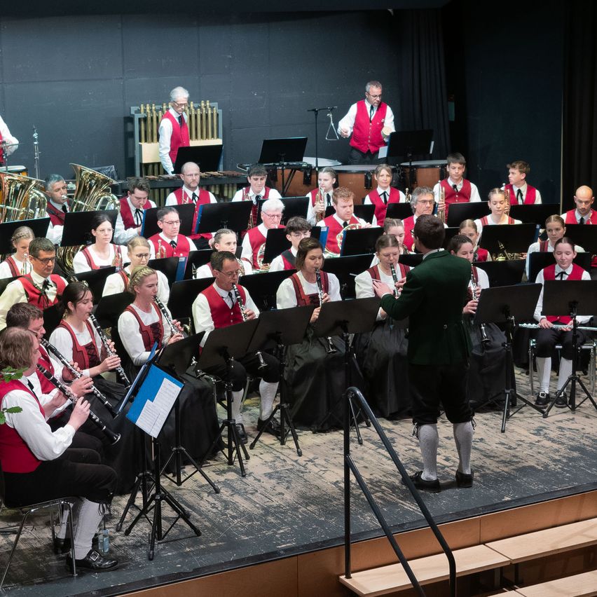 Ein großes Orchester auf der Bühne, Musiker in roten Westen, sitzend und stehend, spielen verschiedene Instrumente, und ein Dirigent in Grün.