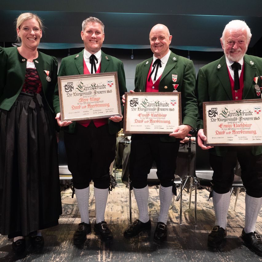 Drei Männer und eine Frau in grünen Jacken und Medaillen stehen auf einer Bühne, lächeln und posieren mit Urkunden. Der Mann auf der linken Seite hält eine Urkunde für Steve Mayer, und der Mann in der Mitte hält eine Urkunde für Ernst Wachsbur. Der Mann auf der rechten Seite hält eine Urkunde für Ernst Nachbur.