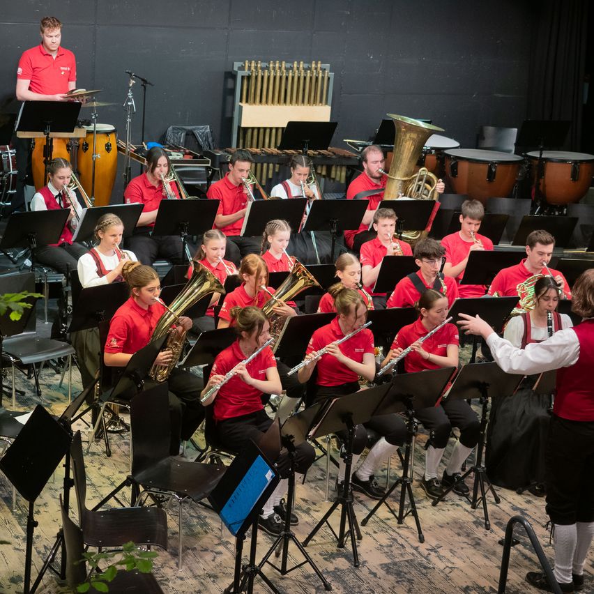 Eine Schulband probt auf einer Bühne mit verschiedenen Instrumenten, darunter Trompeten, Posaunen, Tuben und Trommeln. Die Schüler tragen rote Hemden, einige haben weiße Westen. Der Dirigent steht vorne, und eine Frau in der vorderen Reihe hält ein Notenblatt.