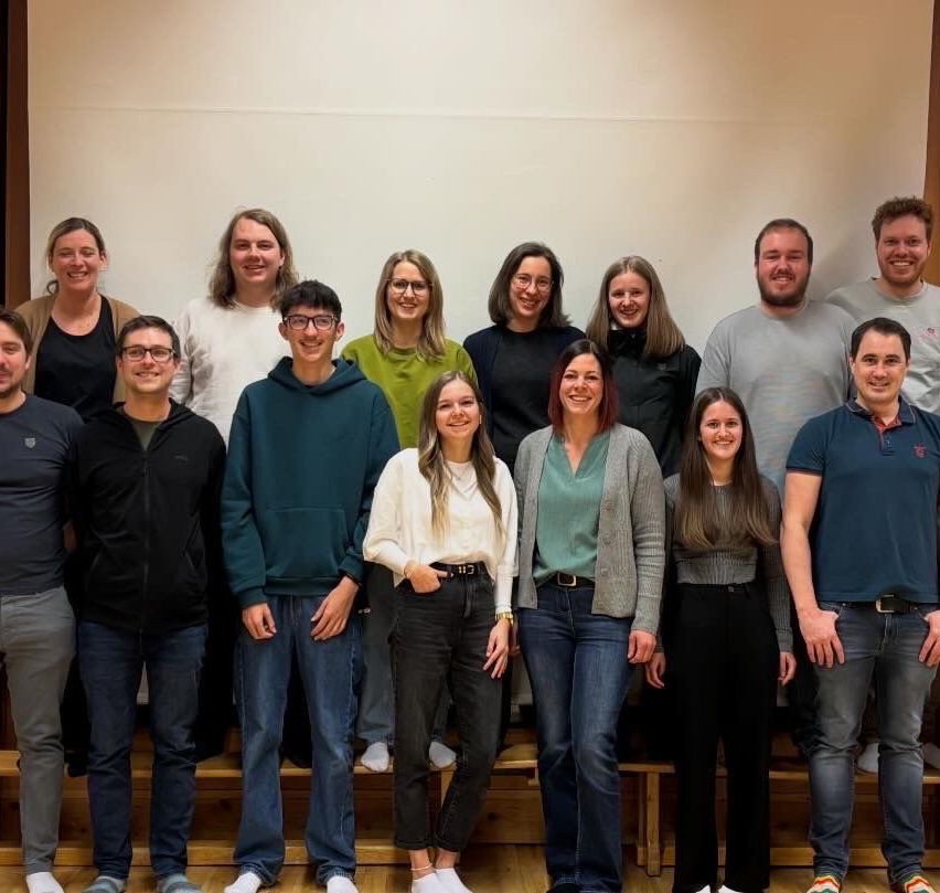 Eine Gruppe junger Menschen und Erwachsener steht lächelnd in einem Raum mit Holzboden und weißer Wand.